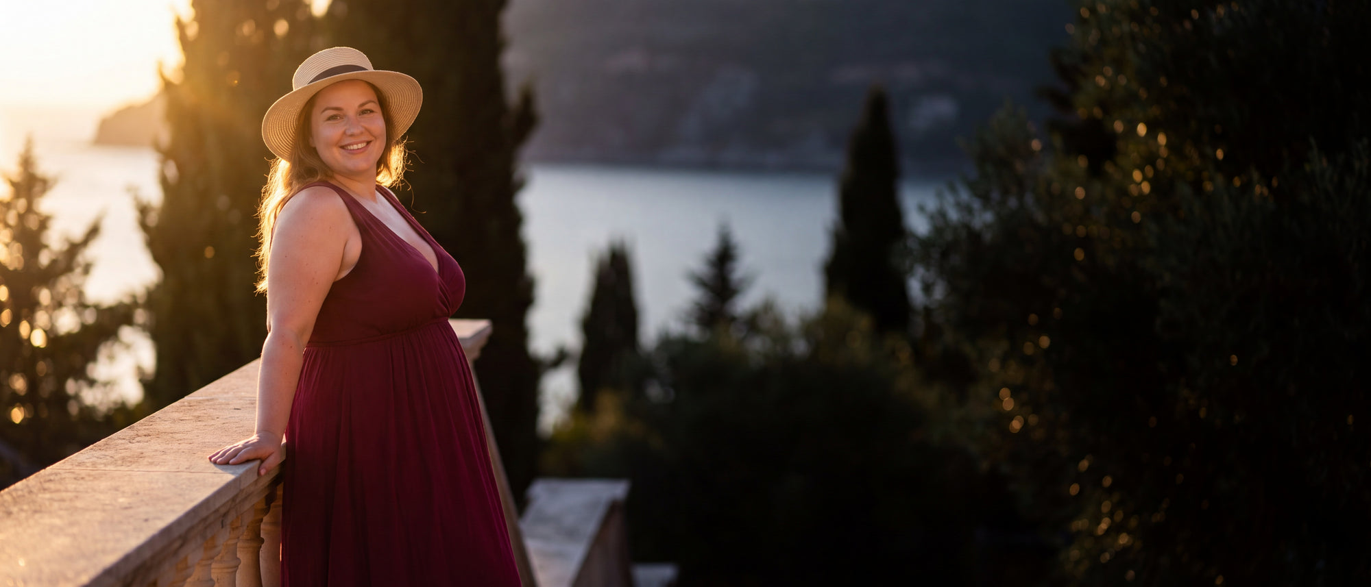Curvy Frau in weinrotem Sommerkleid mit Strohhut auf einer mediterranen Terrasse am Meer im warmen Abendlicht – Betriebsurlaub bei CurvyRausch [KI]