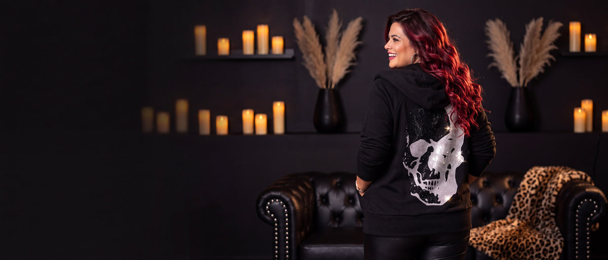 Curvy Frau mit langen weinroten Locken von hinten in schwarzer Sweatjacke mit großem Strass-Skull auf dem Rücken, lachend über die Schulter blickend, in einer kerzenbeleuchteten Boutique – rockige Mode in großen Größen bei CurvyRausch [KI]